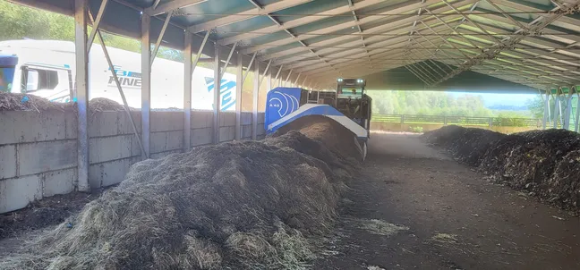 Notranjost pokritega skladiščnega prostora z odprtimi stranicami. Na tleh so dolge kupe temnega, delno razkrojenega rastlinskega materiala. V sredini stoji kmetijski stroj. Ob straneh so betonski bloki, skozi odprtine pa je videti zeleno drevje in naravo v ozadju.