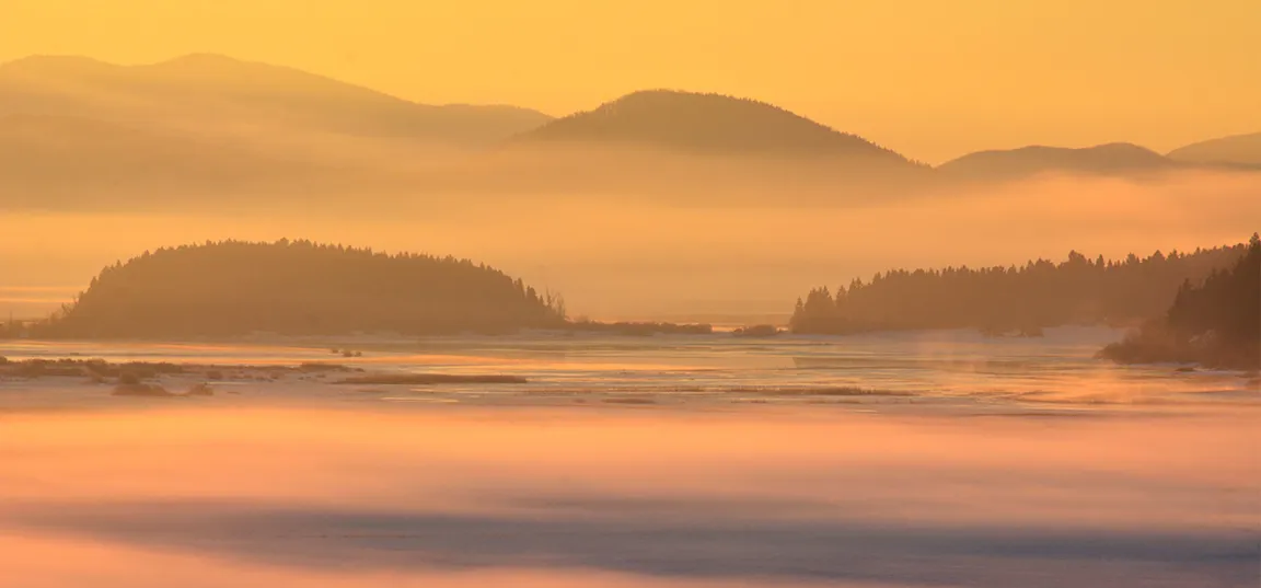 Topel oranžen sončni vzhod nad megleno pokrajino z nežnimi plastmi megle, ki prekrivajo jezero, v ozadju pa so silhuete gričev in gozdnatih otokov.