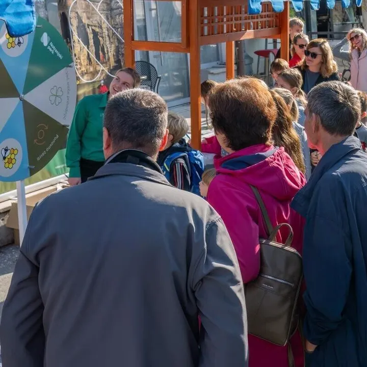 Skupina ljudi na prostem stoji pred kolesom sreče. V ospredju so opazovalci obrnjeni s hrbtom proti kameri, v ozadju pa je sončen dan in nekaj lesenih struktur.