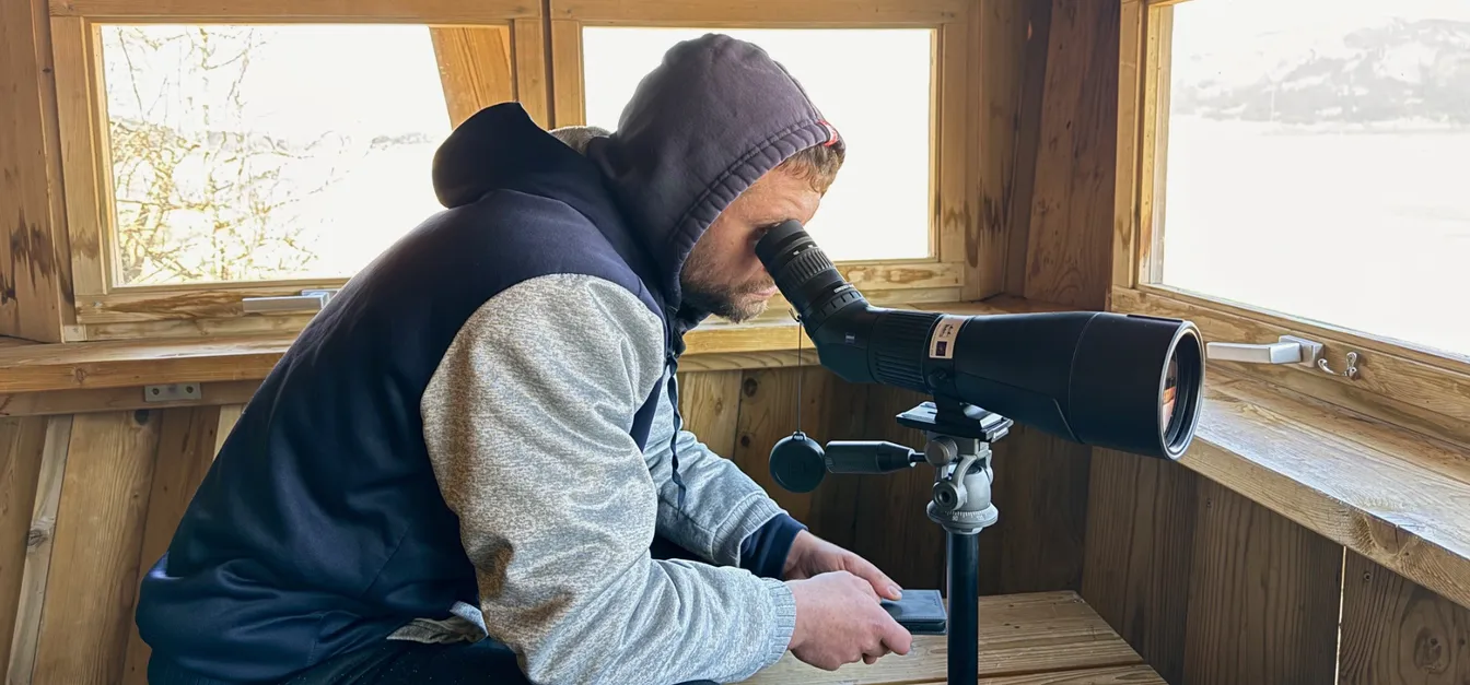 Oseba sedi v leseni opazovalnici in gleda skozi teleskop, nameščen na stojalu; skozi okna opazovalnice je v ozadju vidna naravna krajina.