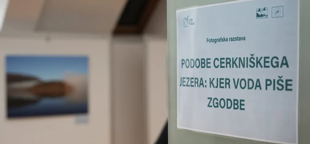 Na desni strani je na steni pritrjen natisnjen plakat z napisom »Fotografska razstava« in naslovom »Podobe Cerkniškega jezera: kjer voda piše zgodbe«. V ozadju levo je zamegljena fotografija pokrajine z jezerom v notranjem prostoru razstave.