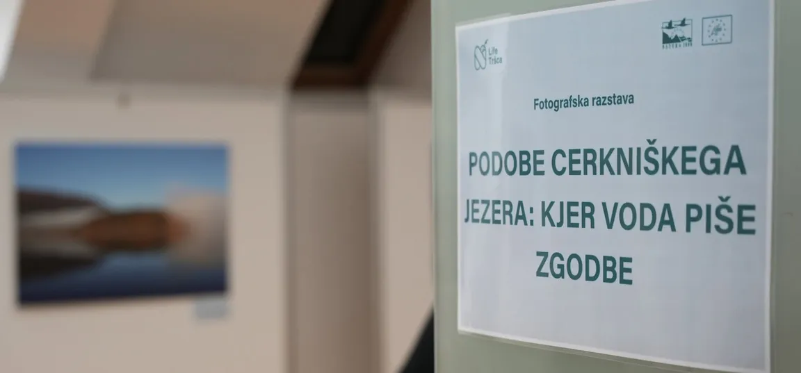 Na desni strani je na steni pritrjen natisnjen plakat z napisom »Fotografska razstava« in naslovom »Podobe Cerkniškega jezera: kjer voda piše zgodbe«. V ozadju levo je zamegljena fotografija pokrajine z jezerom v notranjem prostoru razstave.