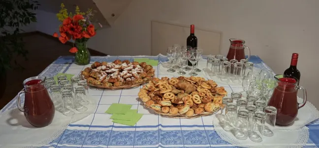 Na mizi s karo prtom so razporejeni krožniki s sladkim pecivom (piškoti in flancati), okoli pa številni prazni kozarci za pijačo. Na levi strani je vrč z rdečim napitkom in šopek rdečih rož, na desni pa steklenica vina ter dodatni vrč z rdečo pijačo.