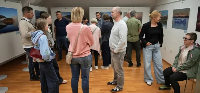 Skupina obiskovalcev stoji in se pogovarja v razstavnem prostoru z lesenim podom. Na stenah so razstavljene fotografije pokrajine, ob robu prostora pa so stoli, na katerih nekaj ljudi sedi. Posamezniki so obrnjeni drug proti drugemu v manjših skupinah, v ospredju je več oseb, oblečenih v sproščena oblačila.