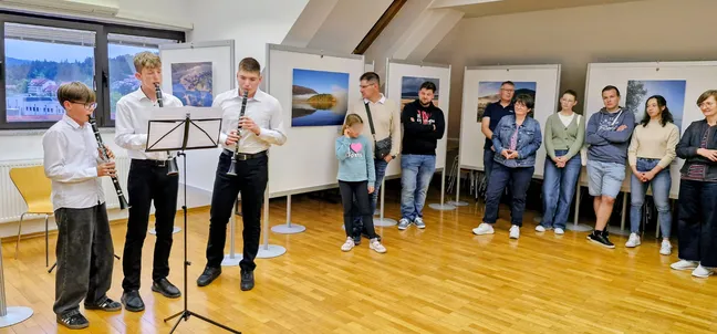V razstavnem prostoru z lesenim podom ob oknu stojijo trije otroci z glasbili (klarinet in saksofon) in igrajo ob notnem stojalu. Ob steni nasproti stoji skupina obiskovalcev, ki opazujejo nastop, na stenah pa so razstavljene fotografije pokrajine.