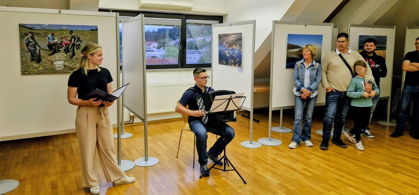 V razstavnem prostoru z lesenim podom sedi oseba na stolu z glasbilom (harmonika) za notnim stojalom, ob strani stoji oseba z listi v roki. Ob desni steni stoji skupina obiskovalcev, na stenah pa so razstavljene fotografije pokrajine.