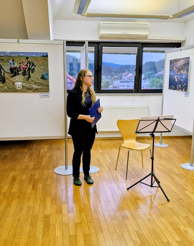 V razstavnem prostoru z lesenim podom stoji oseba z listi v roki ob notnem stojalu. Za osebo je veliko okno z razgledom, na stenah pa so razstavljene fotografije. V prostoru je tudi lesen stol, postavljen ob steni.