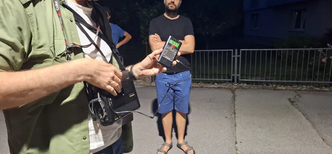 Na večernem terenskem opazovanju oseba v ospredju drži ultrazvočni detektor za poslušanje netopirjev. Ob detektorju visi torba za opremo, v ozadju pa ob kovinski ograji stoji še ena oseba. Prizor je osvetljen z umetno svetlobo, za ograjo je viden del stavbe.