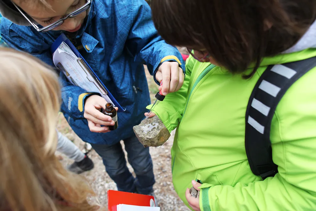 Children exploring rocks outdoors – one holding a rock, the other a bottle with tincture; outdoor classroom, nature day, learning in nature.