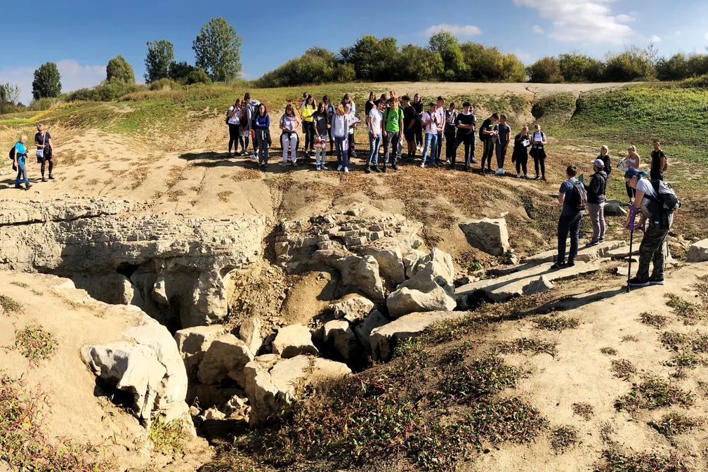 Students are standing on the dry intermmitent Lake Cerknica, where sinkholes through which the water drains are visible. In the background, there are trees standing along the edge of the lake.