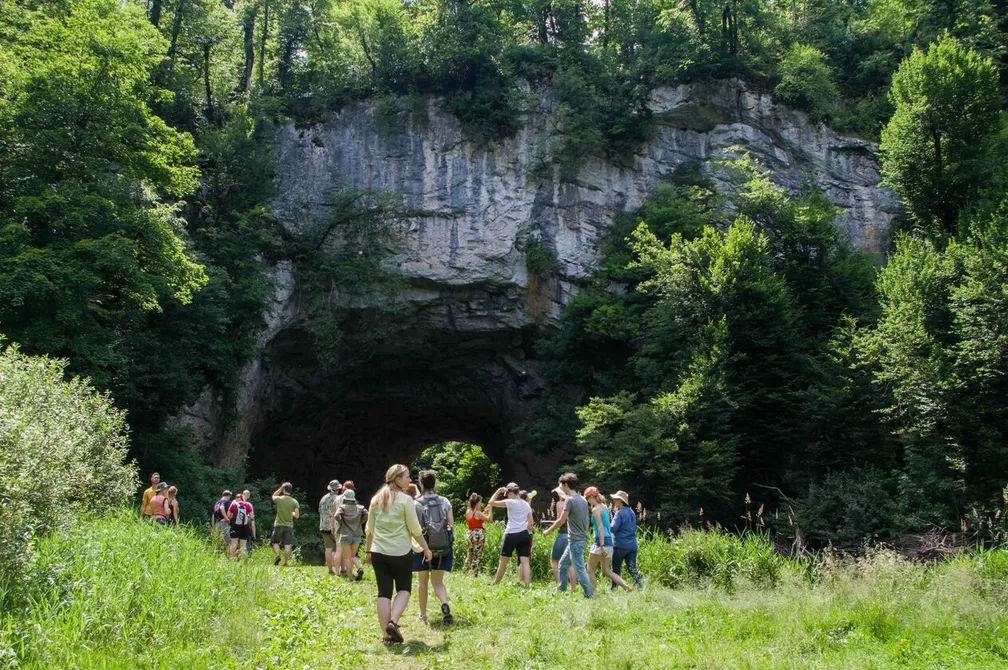 A group of students stands in front of the Big Natural Bridge (Veliki naravni most) in Rakov Škocjan, surrounded by greenery and rocks.