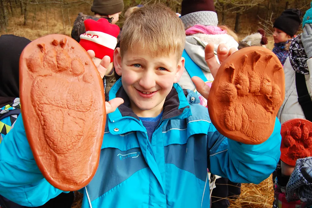 A child in a blue jacket holds up two large clay imprints of animal tracks and smiles; in the background are other people and also holding clay imprints, the scene takes place outdoors in nature.