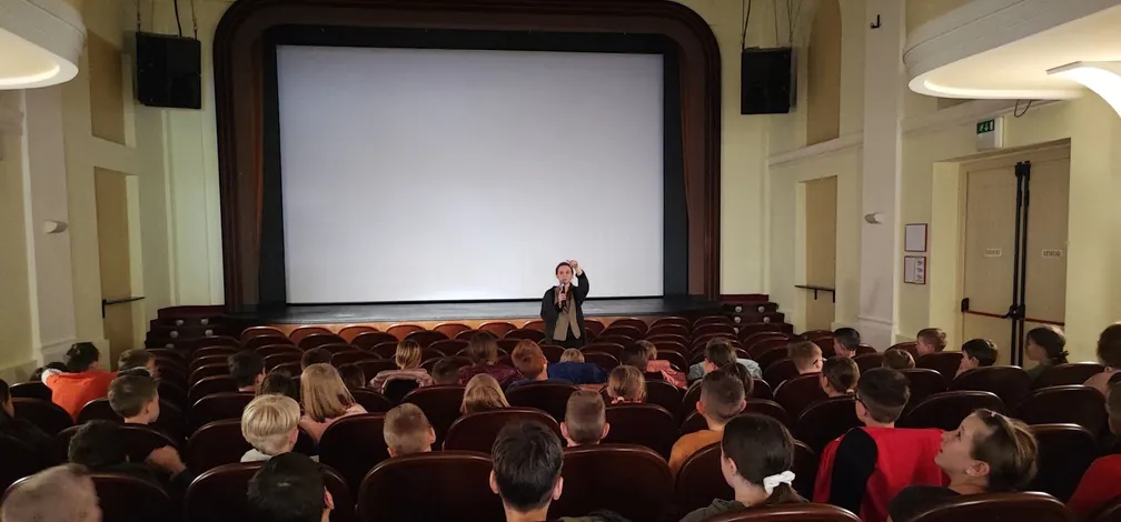 Children seated in a theater space at Cerknica Cultural Center, listening to a speaker standing in front of a large blank screen.