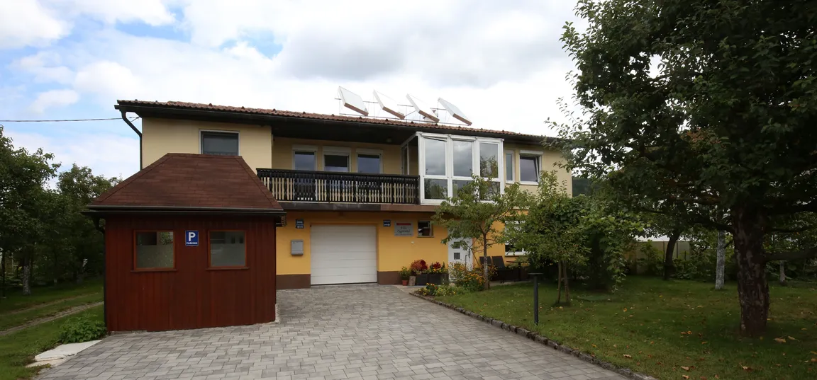 Apartment Vila Zigmund-Two-story house with a yellow facade, brown roof, and a garage with white doors. Solar panels are installed on the roof, the driveway is paved, and the front yard features grass, trees, and bushes.