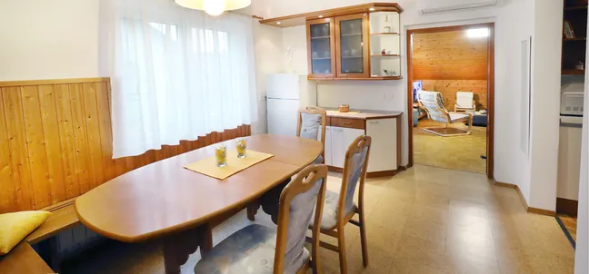 Apartment Vila Zigmund-Dining area with a wooden table, chairs, and a cushioned bench along the wall, decorated with three candles. In the background, a kitchen with cabinets, refrigerator, and counter space, and a living room with wooden walls and furniture.