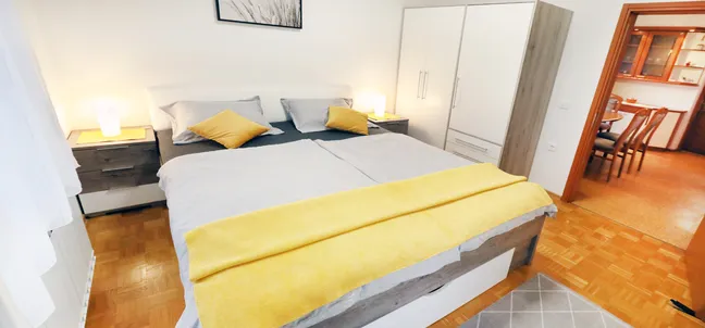 Apartment Vila Zigmund-Bedroom with a large bed, gray and white bedding, yellow accent pillows, and a yellow throw blanket. Bedside tables with lamps flank the bed, and a wardrobe stands on the right. The room features wooden flooring and natural light from the left.