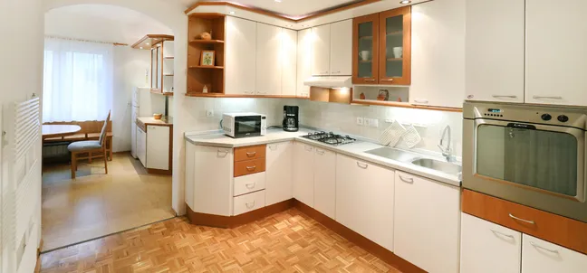 Apartment Vila Zigmund-Modern kitchen with white cabinets and wooden accents, featuring a built-in oven, gas stove, microwave, and a dining area near the window. The parquet flooring and bright layout enhance functionality and style.