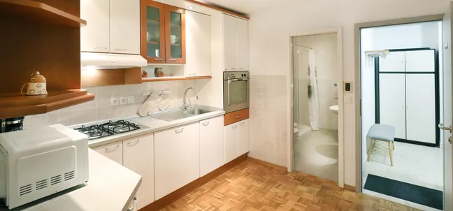 Apartment Vila Zigmund-Kitchen with white cabinets and wooden accents, gas stove, microwave, oven, and parquet flooring. On the right, an open doorway leads to a tiled room with chairs and storage units.