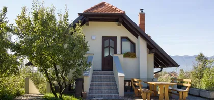 House, where the apartment Knap is, with a red-tiled roof and beige walls, front entrance with steps and wooden door with glass panels, small balcony, flower box by the window, chimney, outdoor seating area, surrounded by greenery and mountains in the background.