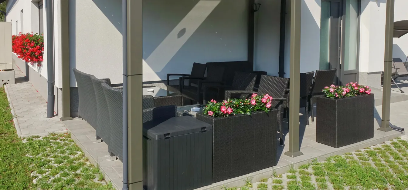 Apartments Anita-Black outdoor seating with flower beds along the edge of the covered terrace. The surrounding area is paved with paving stones, over which grass grows.