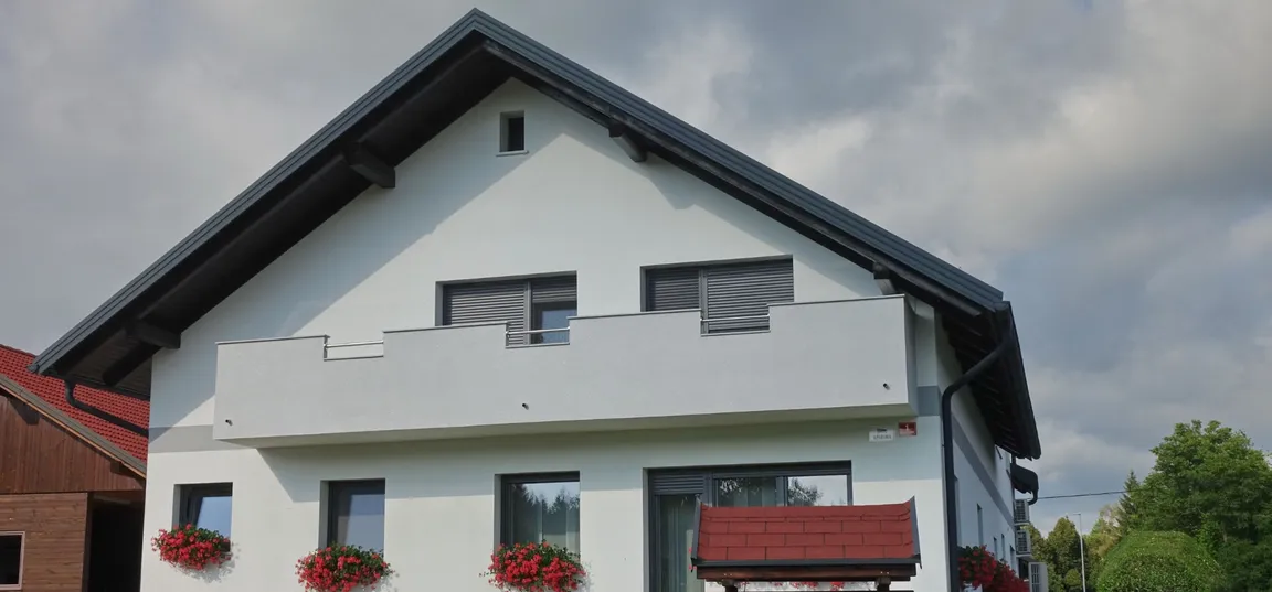 Apartments Anita-Modern two-story house with a white facade, gable roof, and a balcony with a railing. Some windows have flower boxes with red flowers. Greenery is visible in the background under a partly cloudy