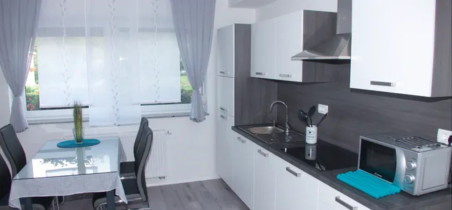 Apartments Anita-Modern kitchen with white cabinets, dark countertops, stainless steel sink, stove with range hood, and a microwave. The dining area features a glass-top table with four chairs and a vase with flowers. Natural light enters through large windows with sheer curtains.