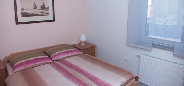 Apartments Anita-Bedroom with a double bed and striped bedding in shades of brown, beige, and pink. Wooden nightstands flank the bed, one with a white lamp. A framed picture of an old town scene hangs above the bed, and a window with sheer curtains and a radiator is on the right.