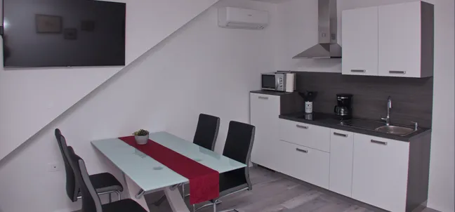 Apartments Anita-Modern kitchen with white cabinets, stainless steel range hood, microwave, coffee maker, and sink. The dining area features a glass-top table with four black chairs and a red table runner. An air conditioner and flat-screen TV are mounted on the walls.
