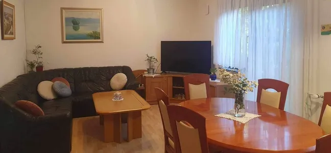 Green Garden Home-vetla dnevna soba z veliko okni, črnim kotnim kavčem, leseno klubsko mizico in jedilno mizo z vazo rož.