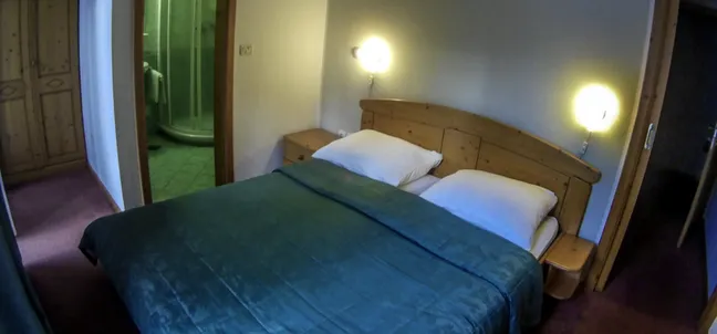 Hotel Rakov Škocjan - double room, bed covered with a green blanket and white pillows, bedside lights on. The bathroom can be seen from the room.