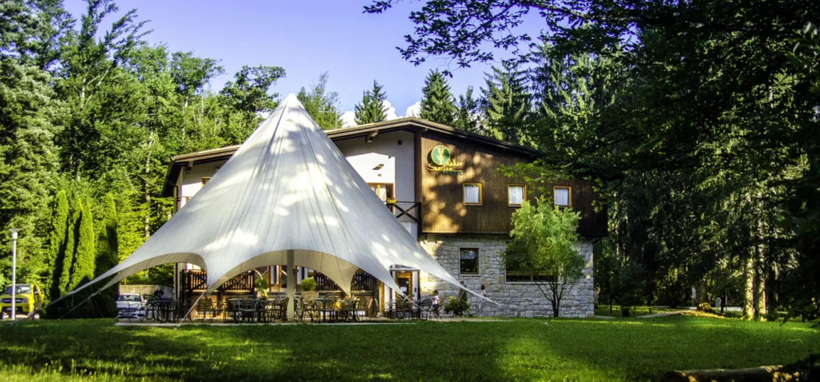 Hotel Rakov Škocjan-A building with a white event tent and an outdoor area with tables and chairs, surrounded by spruce trees and a large garden.