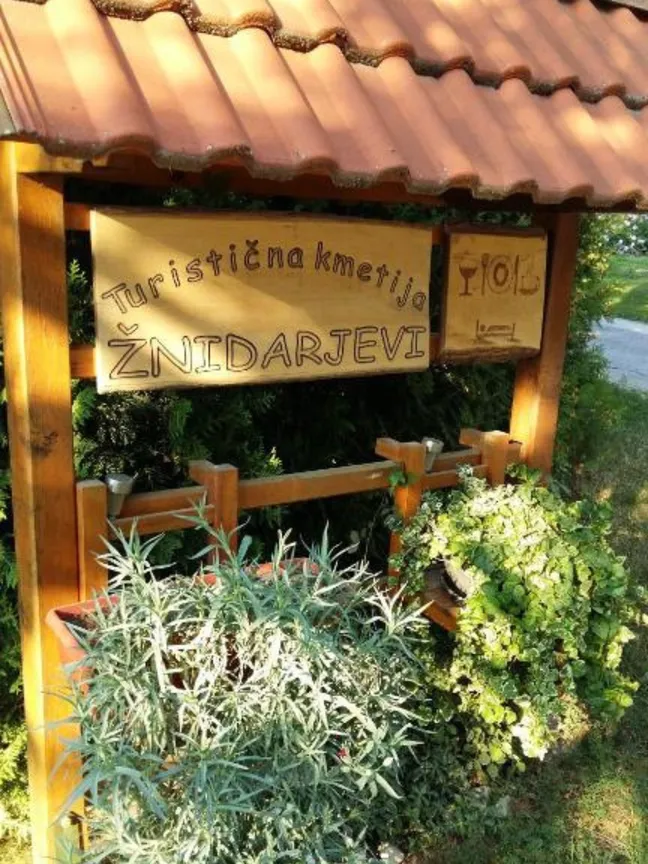 Žnidarjevi Farm-Wooden signboard with tiled roof reading 'Turistična kmetija Žnidarjevi' and 'TIC', surrounded by lush greenery at a Slovenian tourist farm.