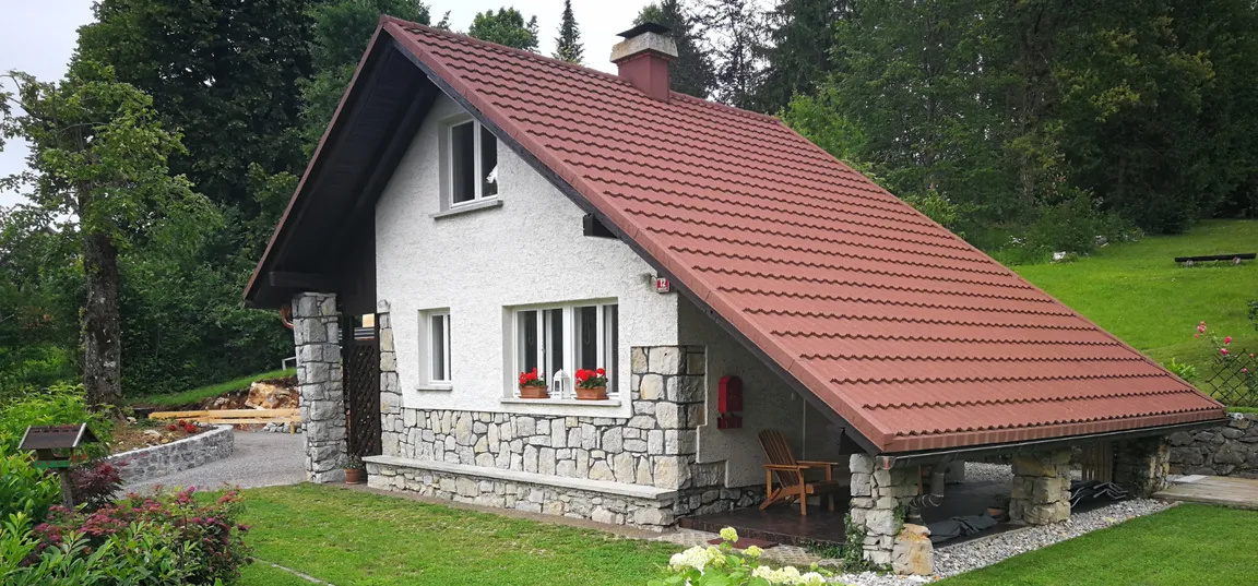 Lake view holiday home-Majhna podeželska hiša z rdečo streho, belimi stenami in kamnitim podnožjem, obdana z zelenjem in cvetličnimi okenskimi koriti.