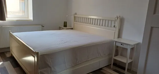 Layka flat-Spalnica z veliko posteljo z belim okvirjem, nočnima omaricama in zeleno svetilko, naravno svetlobo iz okna ter lesenim podom – svetel in udoben prostor za počitek.