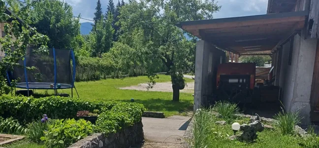 Layka flat-Zadnje dvorišče z zelenico, trampolinom, potko in pokritim prostorom s traktorjem – mirno podeželsko okolje z drevesi in pogledom na gore.