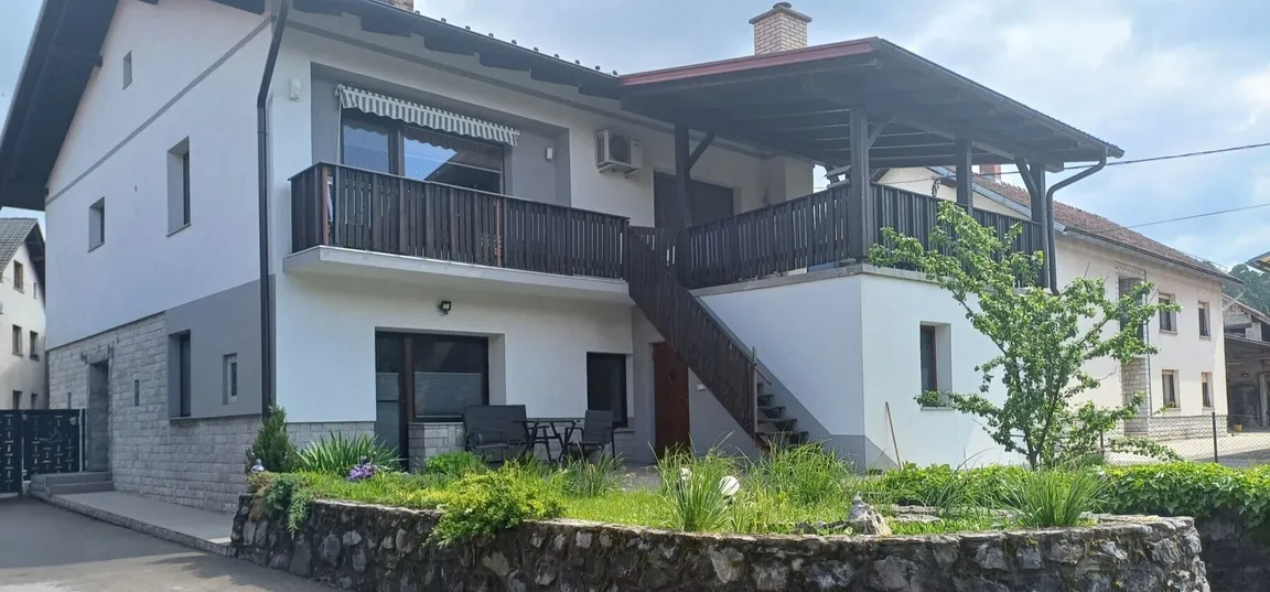 Layka flat-Dvonadstropna hiša z belo fasado, lesenim balkonom in zunanjimi stopnicami, obdana s kamnitim opornim zidom in zelenjem, v ozadju so še druge hiše.