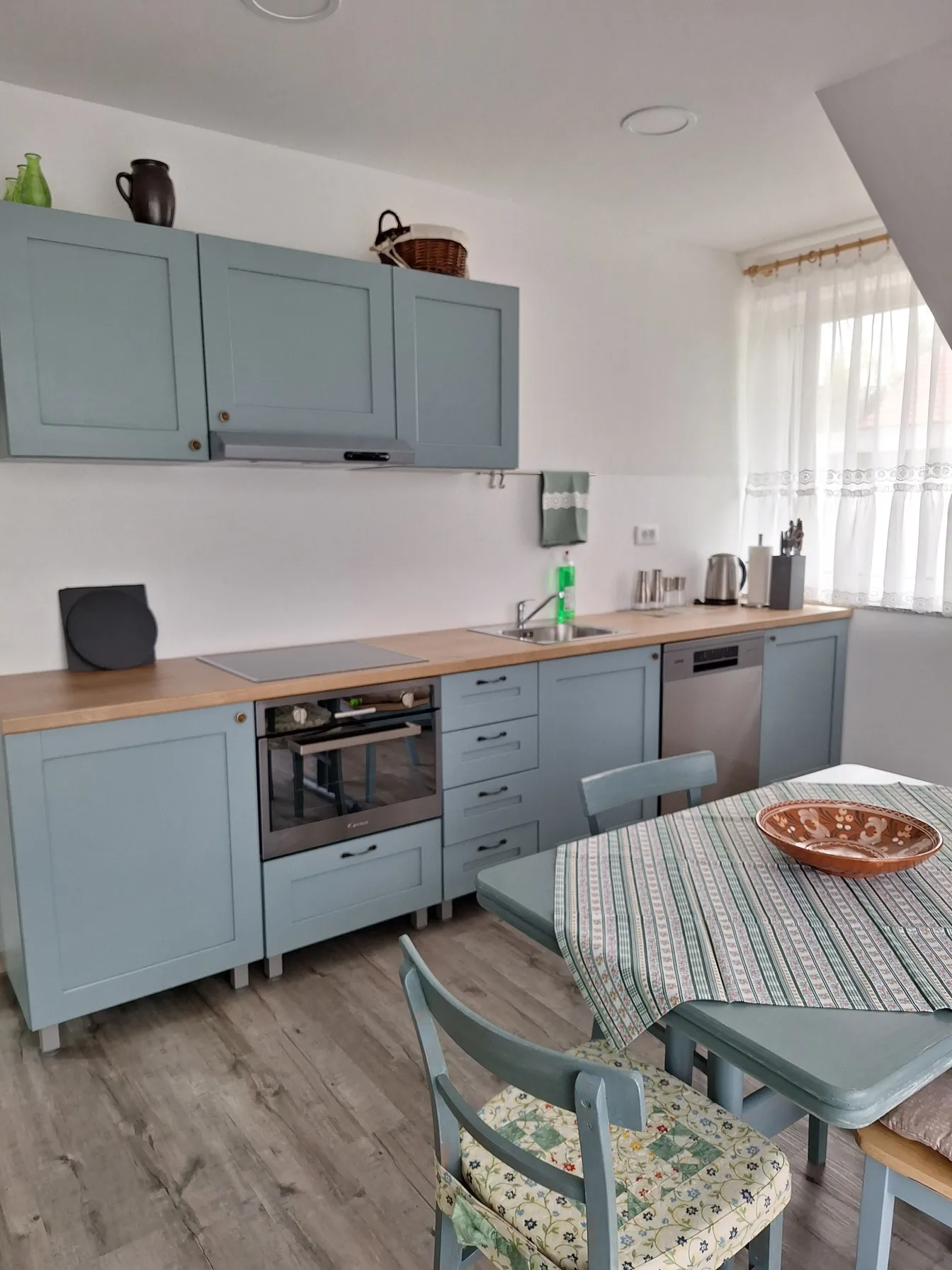 Matejin apartma-sodobna kuhinja s svetlo modrimi omaricami, lesenim pultom, vgrajeno pečico in pomivalnim strojem ter jedilno mizo s prtom – svetel in urejen kuhinjski prostor.