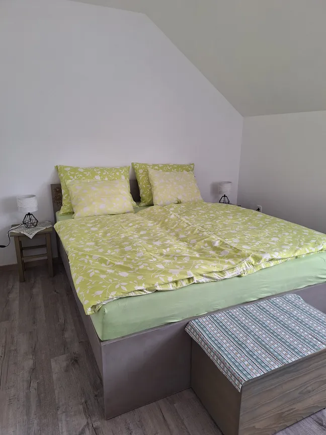 Matejin apartma- Spalnica z urejeno posteljo in svetlo zeleno-belo posteljnino, dvema nočnima omaricama z lučkami ter klopjo z modro-belim vzorcem.