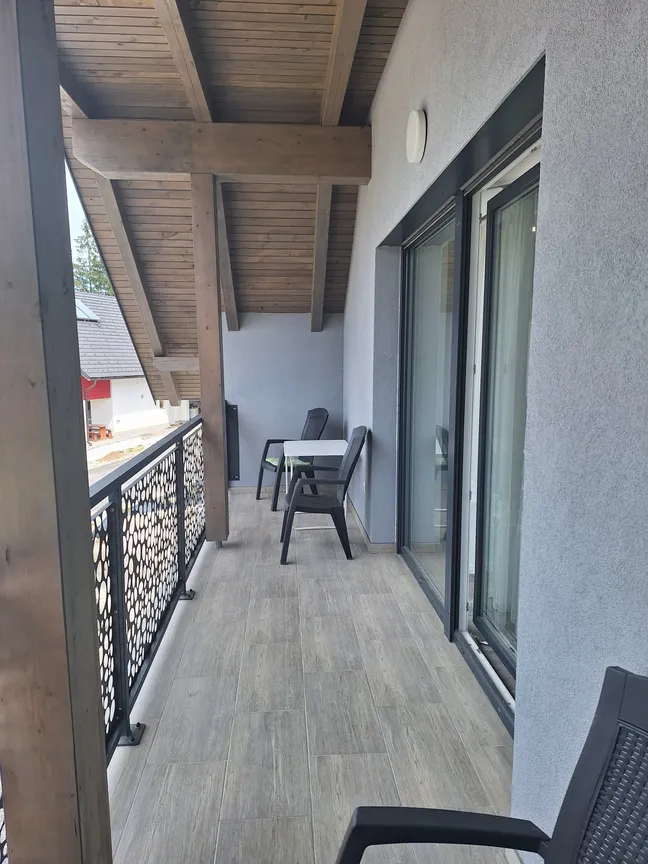 Matejin apartma-balkon z lesenimi tlemi in stropom, dvema črnima stoloma in mizico ob stekleni steni.