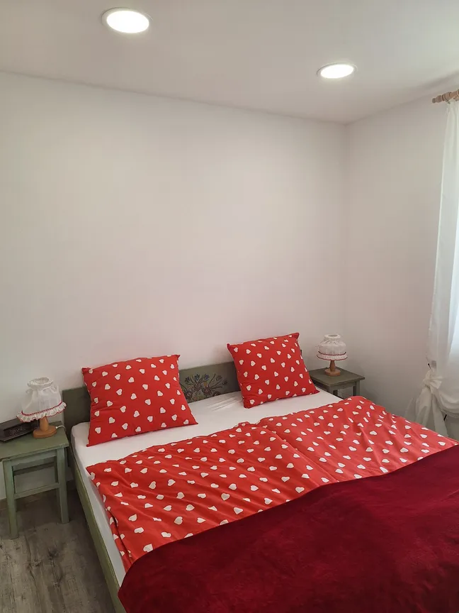 Matejin apartma-Spalnica z rdečo posteljnino z belimi srčki, dvema nočnima omaricama z lučkami, belimi zavesami in lesenim podom.