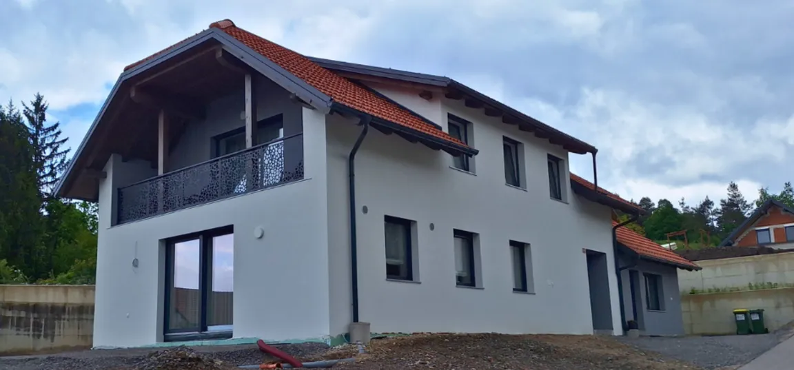 Matejin apartma-Sodobna dvonadstropna hiša z belo fasado in rdečo streho, balkonom z okrasno ograjo ter okolico v fazi urejanja.