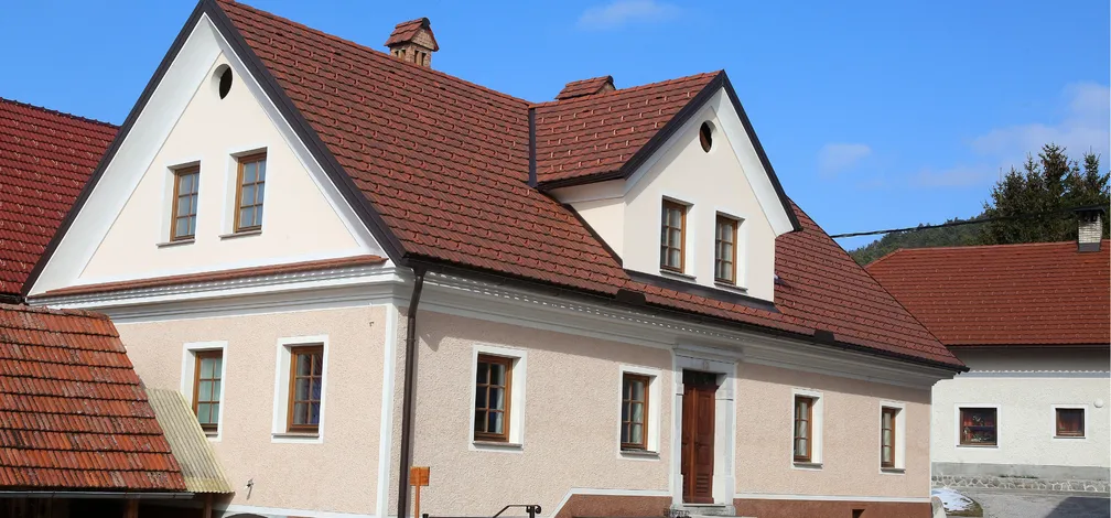 Notranjska hiša-Dvonadstropna hiša z rdečo streho, bež fasado in belimi okenskimi okvirji – tradicionalna arhitektura z lesenimi vrati in trikotnimi čelami v podeželskem okolju.