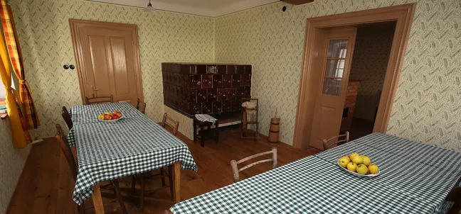 Notranjska hiša-Traditional dining room with wooden flooring, two tables covered with green and white checkered tablecloths, and bowls of fruit. The room features a tiled stove, wooden chairs, yellow curtains, and both open and closed doors leading to other rooms.