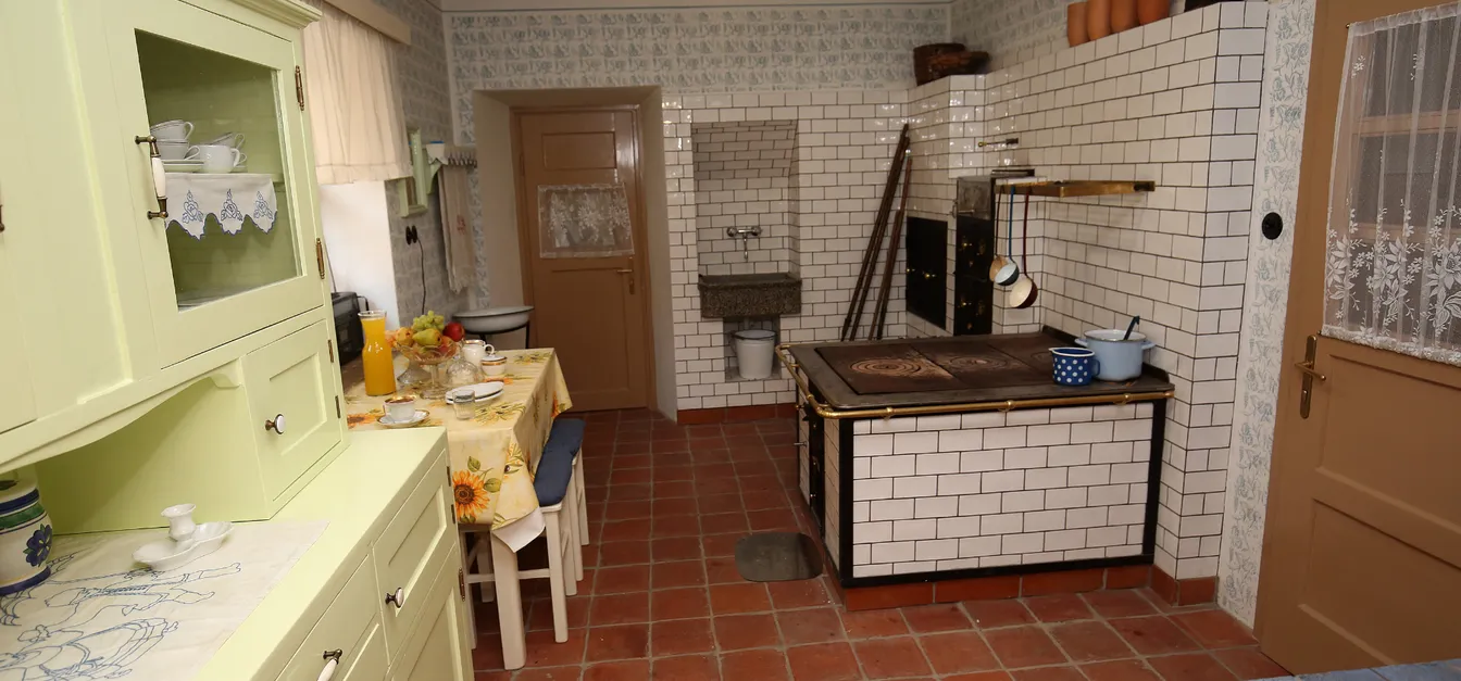 Notranjska hiša-Vintage kitchen with tiled stove, green wooden cabinet, and table with floral tablecloth set for breakfast. There are pots on the stove and ladles hanging above it.