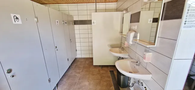 Kamp in PZA - Kmetija z nastanitvijo Tekavča ograda-Javna sanitarna enota z več WC kabinami na levi strani, označenimi s piktogrami. Na desni strani sta dve umivalniški enoti z ogledali, milniki in sušilcem za roke. Stene so obložene s keramičnimi ploščicami v belih in sivih odtenkih, tla pa so rjavo-sive barve. Na stropu je lesena obloga, kar daje prostoru topel videz.