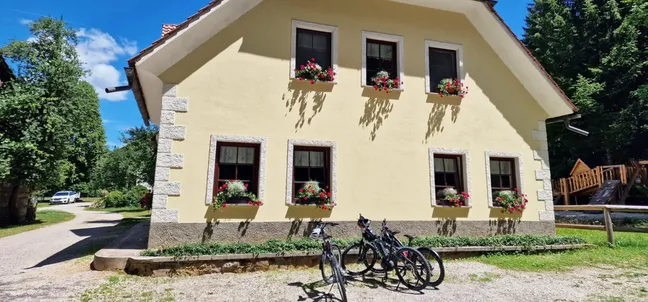 Kamp in PZA - Kmetija z nastanitvijo Tekavča ograda- Rumena podeželska hiša z rdečo streho in šestimi okni, okrašenimi z rdečimi cvetličnimi koriti. Hiša stoji ob makadamski poti, v ozadju so drevesa in lesena terasa. Pred hišo so parkirana tri gorska kolesa na travnati površini. Nebo je jasno modro, okolica je zelena in naravna.