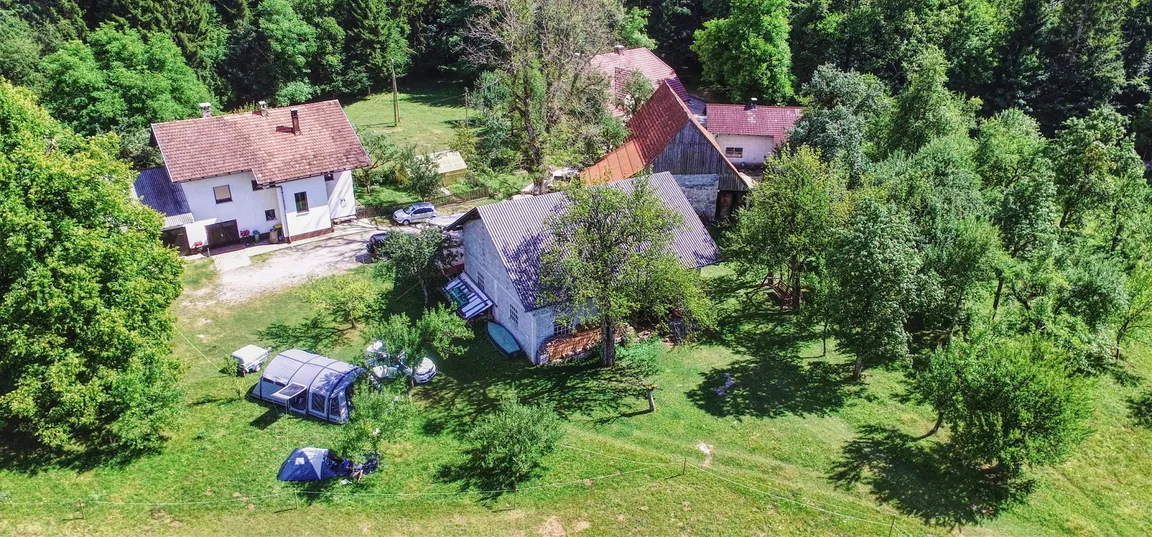 Kamp in PZA - Kmetija z nastanitvijo Tekavča ograda-Posnetek iz zraka, ki prikazuje več hiš na podeželju, obdane z zelenimi drevesi in travniki. V ospredju je večja hiša z rdečo streho, poleg nje manjša stavba in nekaj pomožnih objektov. Na travniku pred hišami so postavljeni šotori in prikolica, kar nakazuje na kampiranje. Okolica je naravna, z veliko zelenja in gozdnim robom v ozadju.