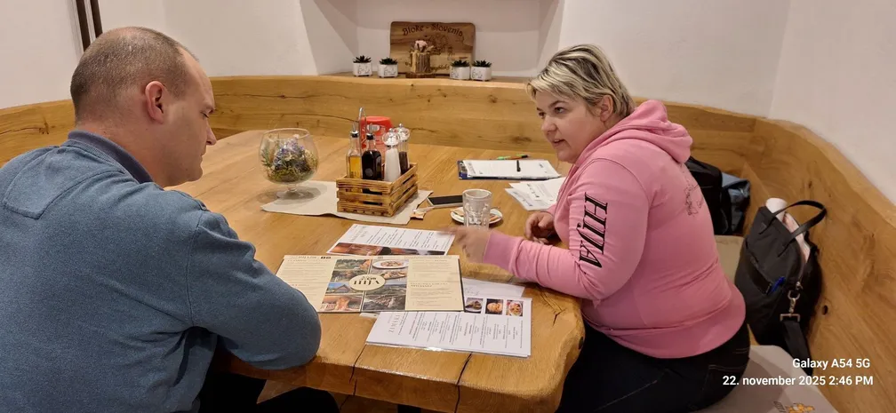 Moški in ženska sedita ob mizi, na kateri so zloženi jedilniki, ter se pogovarjata. Miza in klopi ob njej so iz masivnega lesa.