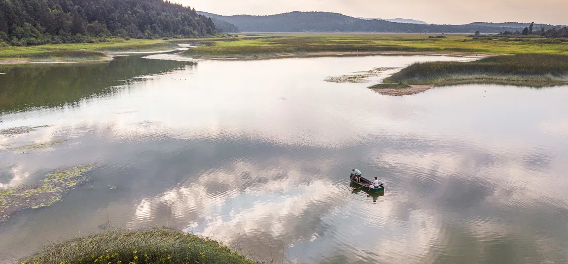 Pogled iz zraka na mirno jezero, obdano z zelenimi močvirnatimi travniki. Na sredini jezera je majhen čoln z dvema osebama, ki veslata po mirni vodni površini, na kateri se odražajo oblaki.