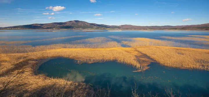 Mirna pokrajina z velikim jezerom, obdanem z visokimi zlatorjavimi trstičji. V ozadju se raztezajo griči in gore pod modrim nebom z razpršenimi oblaki. Voda odseva nebo in okoliško rastlinje ter ustvarja spokojno in slikovito vzdušje.